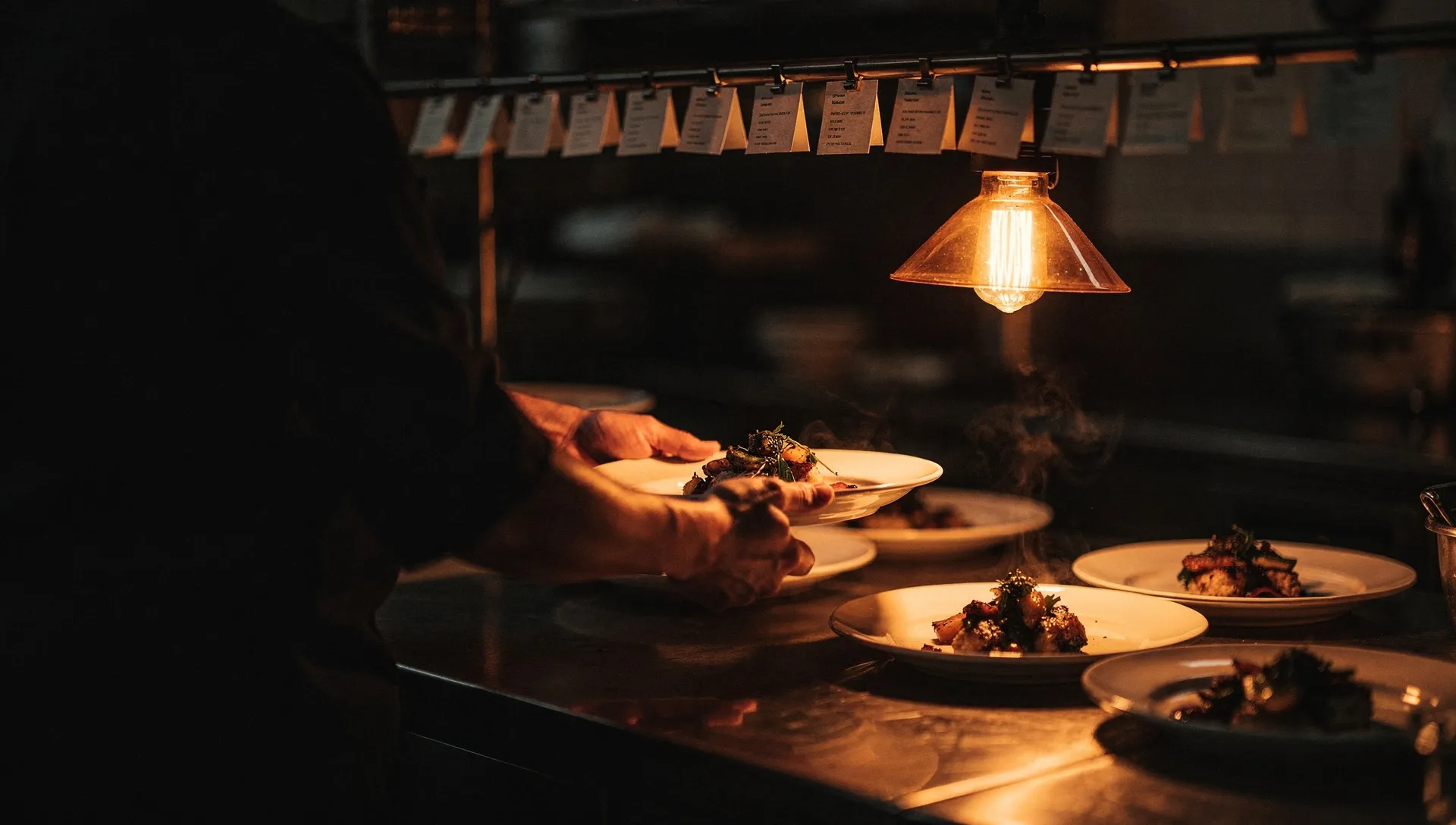 A restaurant kitchen pass at evening service, plated dishes and a rail of order tickets lit by a single warm amber heat lamp